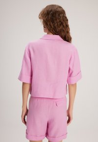 Camicetta rosa in lino a maniche corte con colletto e maniche cuffiate, abbinata a pantaloni corti in lino rosa coordinati con una tasca sul retro.