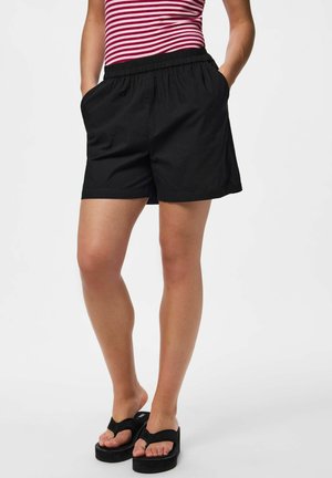 Persoană care poartă pantaloni scurți negri cu talie elastică și papuci negri cu platformă, stând cu mâinile în buzunare, pe un fundal alb.