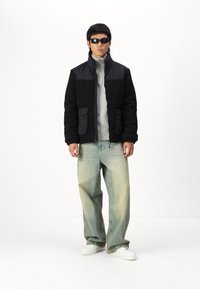 Black and grey fleece jacket with two front pockets, worn over a grey zip-up top. Light blue wide-leg jeans and white sneakers complete the outfit.