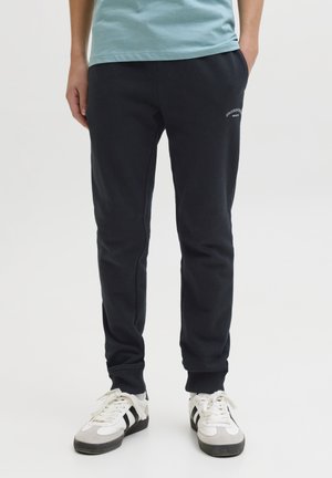 Persona che indossa pantaloni jogger neri con piccolo logo bianco sulla coscia, abbinati a sneakers bianche con strisce nere, in piedi con le mani in tasca.