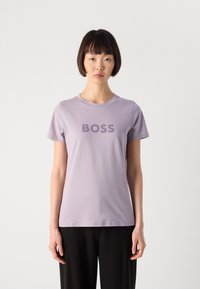 Moteris su trumpais juodais plaukais, vilkinti šviesiai purpurinę marškinėlį su užrašu "BOSS" ir juodas kelnes, stovi prieš paprastą baltą foną.