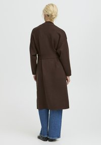 Donna bionda con un raccolto basso che indossa un lungo cappotto marrone scuro con cintura sopra a dei jeans blu e scarpe nere, in piedi con la schiena rivolta in avanti.