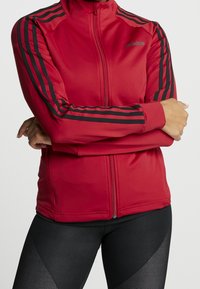 Rote Sportjacke aus dehnbarem Material. Hat schwarze Streifen an den Ärmeln, einen Reißverschluss vorne und eine taillierte Passform.