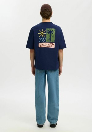 Homme portant un t-shirt bleu marine avec des motifs colorés de soleil, vagues, palmier et silhouette allongée dans le dos, jean bleu et casquette marron.