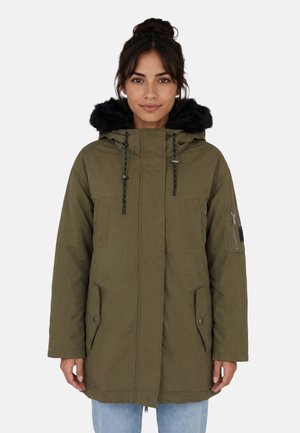 Donna che indossa un parka verde oliva con cappuccio in pelliccia nera e tasca con cerniera sulla manica, in piedi davanti a uno sfondo semplice.