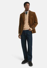 Mann in braunem Cordblazer, beigefarbenem Quarter-Zip-Pullover, blauer Krawatte, dunkelblauen Jeans und schwarzen Schuhen, der mit einer Hand in der Tasche steht.