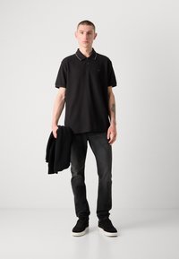 Svart polotröja med vit krage, kombinerad med mörkgrå jeans. Modellen håller en svart tröja och har på sig svarta sneakers med vita sulor.