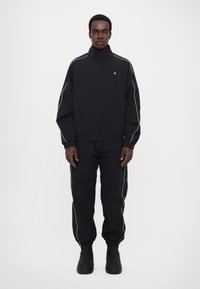 Svart idrottsoverall med en zippered jacka och byxor, med vit piping längs axlarna och sidorna. Slät tygstruktur.