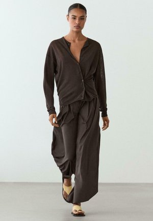 Kobieta spaceruje w pomieszczeniu, nosząc luźną, ciemnobrązową bluzkę zapinaną na guziki oraz pasujące szerokie spodnie i beżowe sandały.