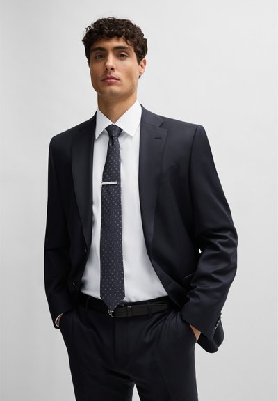 Costume marine avec un fini brillant, chemise blanche et cravate à pois noirs avec une pince à cravate argentée. Design ajusté avec revers crantés.