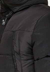 Sort dunjakke med quiltet tekstur. Har en høj krave, lynlåslukning og præget branding langs sidesømmen.