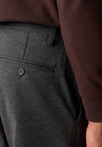 Primer plano de un bolsillo de pantalones gris oscuro con botón y etiqueta, acompañado de la manga de una camisa de manga larga marrón y una mano al lado.