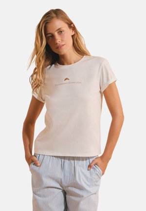 Mujer con cabello largo y rubio que lleva una camiseta blanca con un gráfico de un croissant y texto en francés, combinada con pantalones azul claro a rayas.
