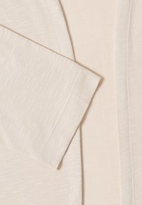 Close-up of a beige knit fabric with a long sleeve and a front edge seam overlap, showing fine texture and stitching details.