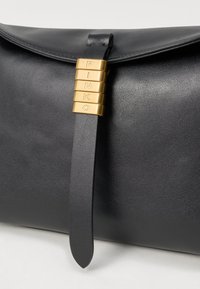 Crna kožna torbica s zaklopom zatvorenim trakicom, na kojoj su zlatne kockice koje vertikalno ispisuju "PINKO".