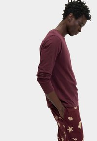 Camiseta de manga larga color burdeos hecha de tela suave, combinada con pantalones burgundy a rayas que presentan detalles dorados de estrellas y símbolos navideños.