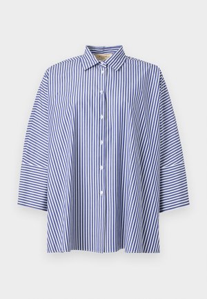 Mujer con cabello largo y oscuro lleva una camisa de rayas azules y blancas, pantalones beige de pierna ancha y chanclas negras, de pie sobre fondo blanco.