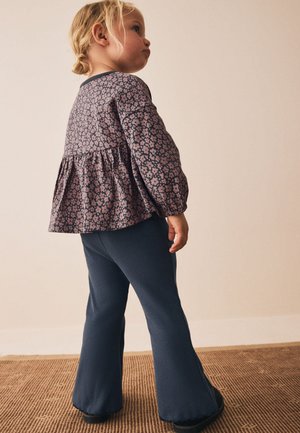 Småbarn med blondt hår iført en blomsterlangærmet top og marineblå vidde bukser står på et tekstureret brunt tæppe mod en ensfarvet væg.