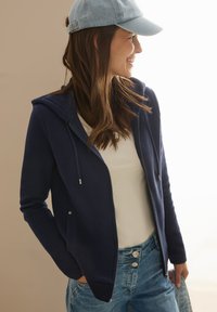 Marinblå zip-up hoodie med en framficka, snörning vid huvan, kombinerad med en vit skjorta och ljusblå denimjeans. Mjuk textur.