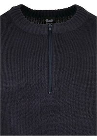 Pletený sveter v tmavo sivej farbe s vysokým golierom, ktorý má štvrtinový zips a textúrované rebrovanie. Štítok obsahuje nápis "Brandit Industrial Wear."