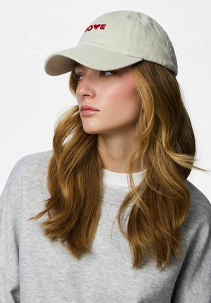 Vrouw met lang golvend lichtbruin haar draagt een lichtgrijze trui en een beige pet met rode tekst "LOVE", kijkt naar links tegen een witte achtergrond.