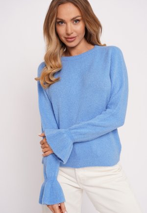 Kobieta o długich blond włosach, ubrana w miękki niebieski sweter z rozszerzanymi mankietami i białe spodnie, pozująca na jasnym, jednolitym tle.