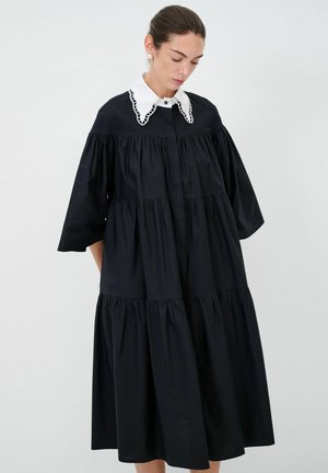 Schwarzes Baumwolklei mit gestufter Gestaltung, versehen mit einem weißen Kragen mit Spitzenbesatz und kurzen, puffärmeligen Ärmeln. Glatte Textur und knielang.