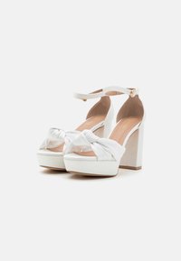 Zapatos de tacón de plataforma blancos con un detalle de nudo en la parte delantera, correa ajustable en el tobillo y un tacón cuadrado y grueso. Material suave y plantilla ligeramente acolchada.