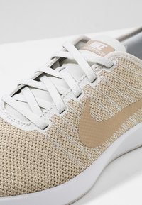 Nike-sneakers med ett ljusbeige nätovanför, vita snören och en beige swoosh-logotyp. Sulan har en strukturerad, böjd design.