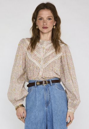 Donna con capelli castani mossi che indossa orecchini a cerchio dorati, una camicetta a maniche lunghe floreale con dettagli in pizzo, jeans blu a vita alta e cintura marrone.