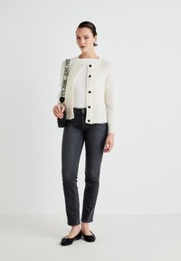White knitted cardigan with round neckline and brown buttons, paired with fitted dark gray jeans and black flats, carrying a black bag.