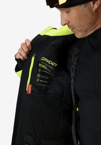Schwarze Skijacke mit neon-grünen Akzenten, ausgestattet mit einer glatten Textur, wasserdichten Etiketten und einer Mesh-Tasche im Innenfutter.