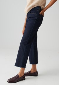 Dark blue cropped jeans with a straight leg, featuring a frayed hem and traditional five-pocket design. Paired with glossy burgundy loafers.