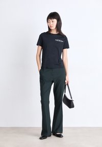 Zwarte katoenen t-shirt met grafische print, gecombineerd met donkergroene broek. Model houdt een zwarte schoudertas vast en staat op een lichte vloer.