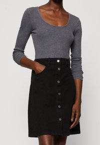 Nainen, joka käyttää tiukasti muotoiltua harmaata pitkähihasta toppia, joka on huhdottu korkeavyötäröiseen mustaan napitettuun hameeseen, jossa on taskut, yksi käsi taskussa.