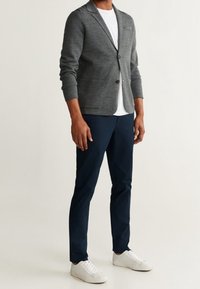 Šedý pletený blazer s dlouhými rukávy přes bílé tričko, spárovaný s námořnickými kalhotami a bílými teniskami. Výrazný detail knoflíku na blazeru.