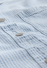 Light blue textured fabric with a grid pattern, featuring metal buttons. Close-up view showing stitching and pocket details.