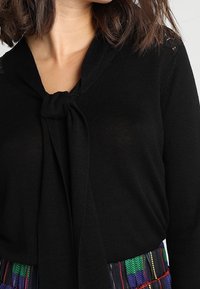 Svart stickad tröja med en knytdetalj vid halsen, som har en mjuk textur och en figurnära design.