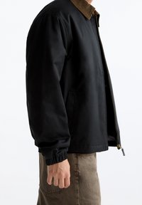 Man in een zwart ritssluitingjack met een bruine ribflonnekraag en een bruine broek, staand tegen een witte achtergrond.