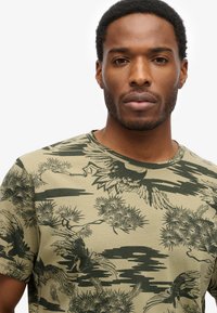 Oliven grøn t-shirt med et tekstureret tryk af sorte fugle og træer. Rund halsudskæring og kortærmede, lavet af letvægts bomuld.