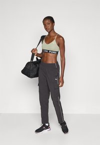 Olīvu zaļš sporta krūšturis ar melnu logotipa lentu, apvienots ar melnām kravas biksēm ar baltām akcentiem un melnām sporta kurpēm. Nesot melnu sporta somu.