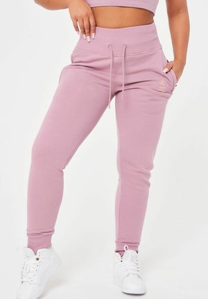 Woman wearing mauve high-waisted joggers with drawstring waist and white sneakers, posing with one hand in pocket and one on hip.