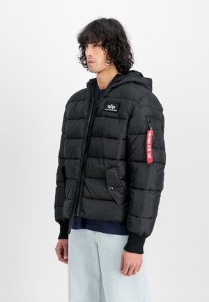 Mann iført en svart pufferjakke med hette, glidelås foran, logo-patch på brystet, og rød merkelapp på ermet, i kombinasjon med lyseblå jeans.