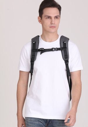 Uomo che indossa una t-shirt bianca e uno zaino con spallacci neri e fibbia sul petto, in piedi davanti a uno sfondo chiaro uniforme.