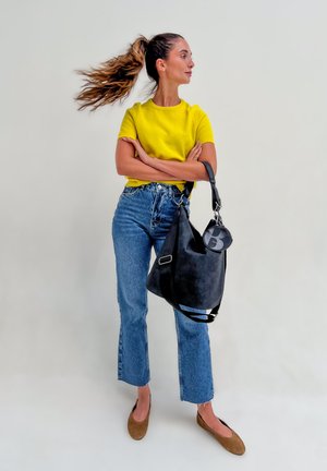 Frau mit langem Zopf, die ein gelbes Oberteil, blaue Jeans, braune Ballerinas trägt und eine große schwarze Handtasche hält, mit verschränkten Armen seitlich stehend.