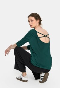 Zielona, luźna bluzka z okrągłym dekoltem i skrzyżowanymi ramiączkami na otwartych plecach, w połączeniu z szerokimi czarnymi spodniami i białymi trampkami.