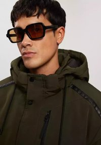 Zielona kurtka z kapturem z kontrastującymi ciemnymi akcentami na ramionach, z zamkiem błyskawicznym w kieszeniach i wysokim kołnierzem. Czarne, oversize'owe okulary przeciwsłoneczne z brązowymi soczewkami.