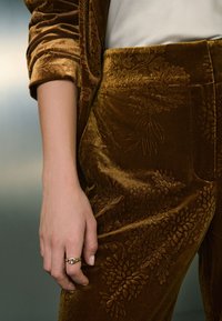 Pantalones de terciopelo en marrón oscuro con relieve floral. La tela tiene una textura suave. Una mano con un anillo decorativo descansa sobre la pretina.