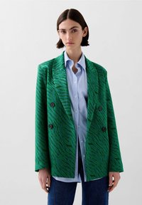 Groene blazer met een tijgerstrippatroon, dubbelzijdig ontwerp, textured stof en zwarte knopen, gedragen over een blauw gestreept overhemd.