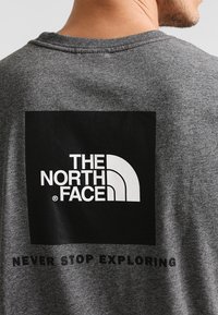 Šedé bavlněné tričko s černým obdélníkovým logem na zadní straně, na kterém je bíle napsáno "THE NORTH FACE" a "NEVER STOP EXPLORING".